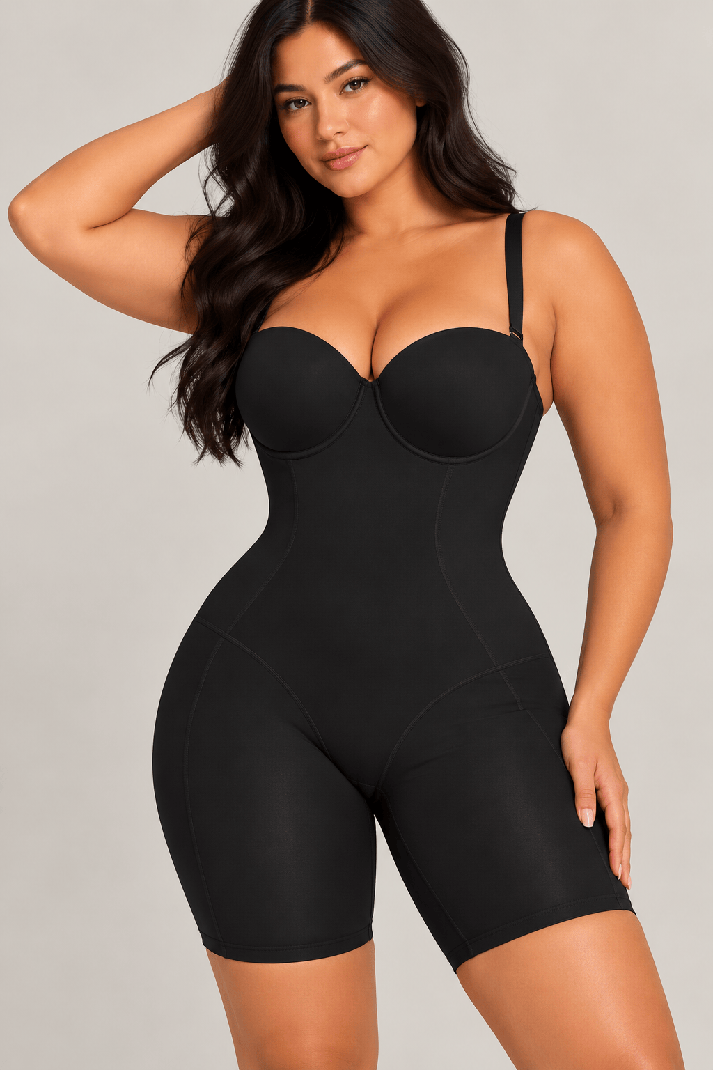 A woman modeling a black cupped mid thigh strapless shapewear bodysuit with a bustier-style upper section and fitted shorts, photographed against a plain light gray studio background.