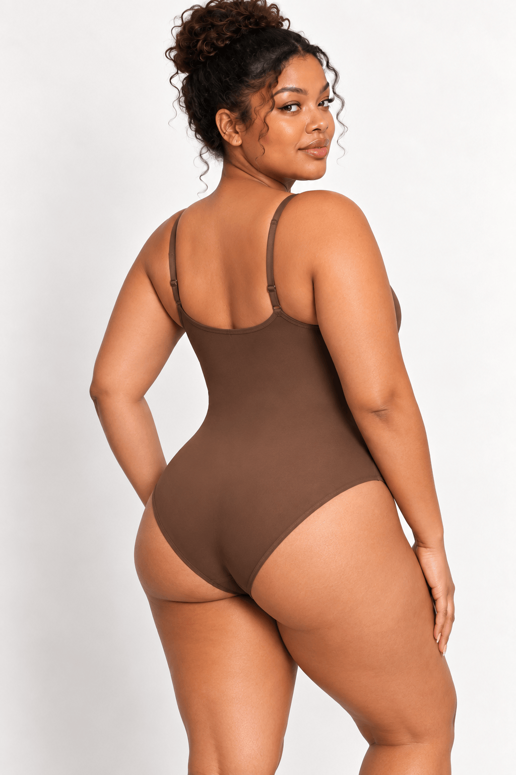 A woman modeling a brown shapewear bodysuit from the back, showing the thin straps and high-cut legs, against a plain light gray background.