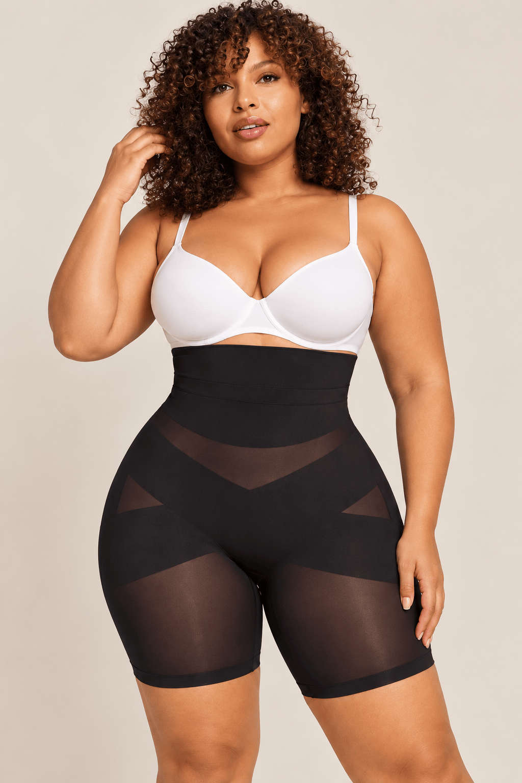 A plus-size model wearing a white bra and black high-waisted shapewear shorts with sheer mesh panels, posed against a solid light beige background.