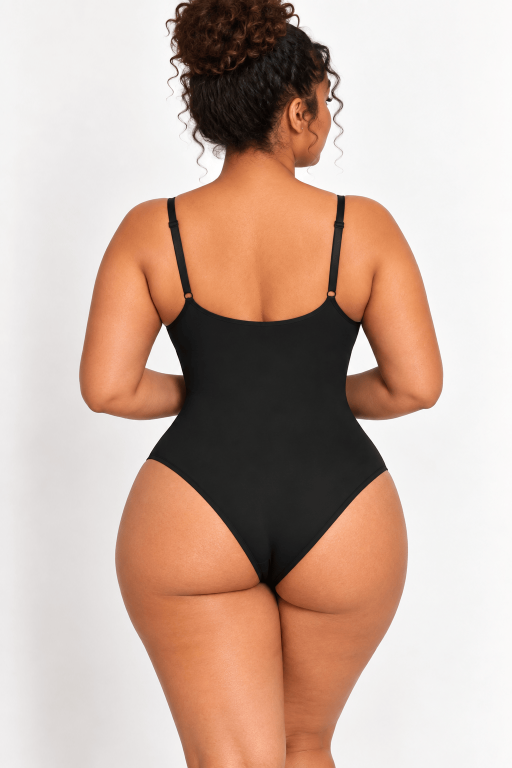 A woman shown from the back wearing a black one-piece bodysuit with thin shoulder straps, photographed against a plain light gray studio background.